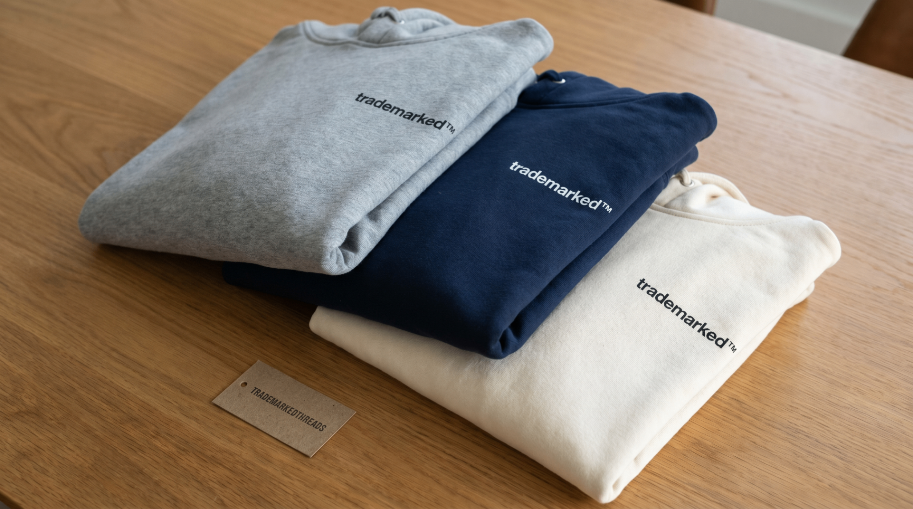 Three folded Trademarked Threads hoodies in heather gray, navy, and ivory on a wood surface.